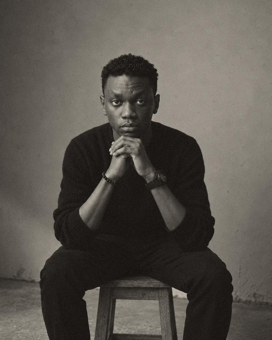 Ahmed Zaki seated on a wooden stool against a bare wall, hands clasped at his chin, in a black knit jumper.